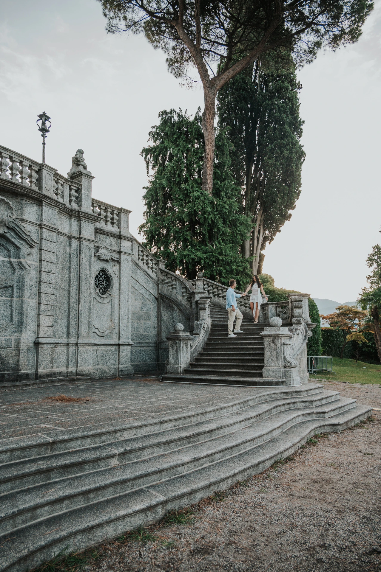 The couple stand on an ornate stone staircase surrounded by tall trees during their Lake Como photoshoot at Parco Civico Teresio Olivelli.