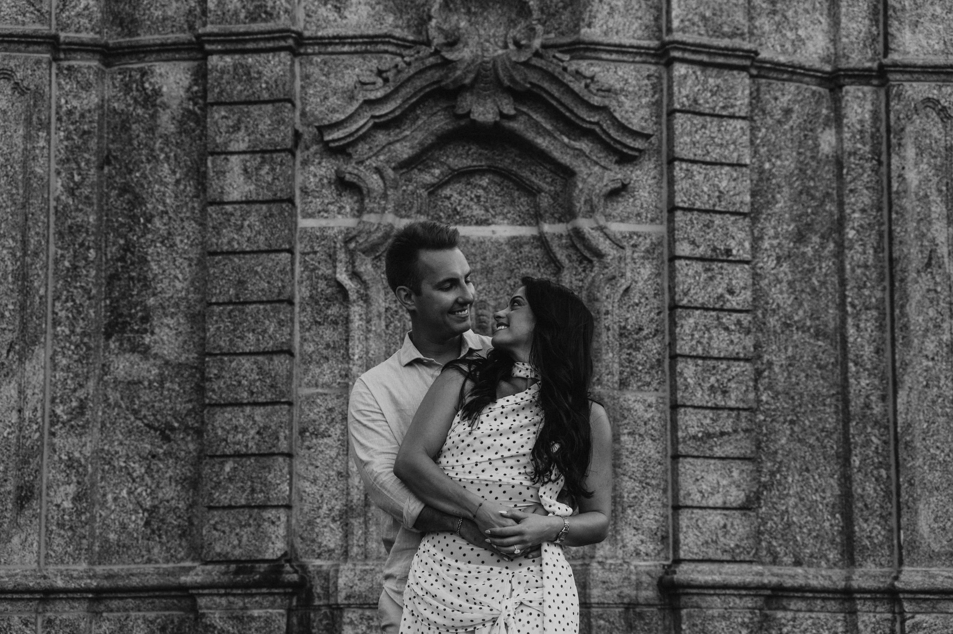 Black-and-white portrait of the couple embracing and looking at each other against carved stone panels during their Lake Como engagement photoshoot.