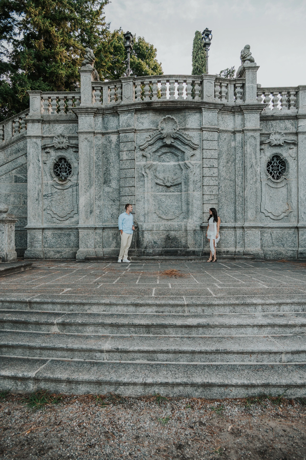 The couple stand apart across the broad stone steps beneath the ornate terrace during their Lake Como photoshoot at Parco Civico Teresio Olivelli.