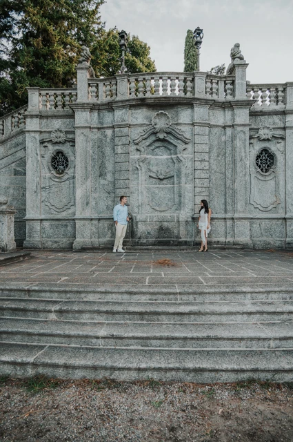 Wide Lake Como photoshoot beneath the park’s ornate stone terrace The couple stand apart across the broad stone steps beneath the ornate terrace during their Lake Como photoshoot at Parco Civico Teresio Olivelli.