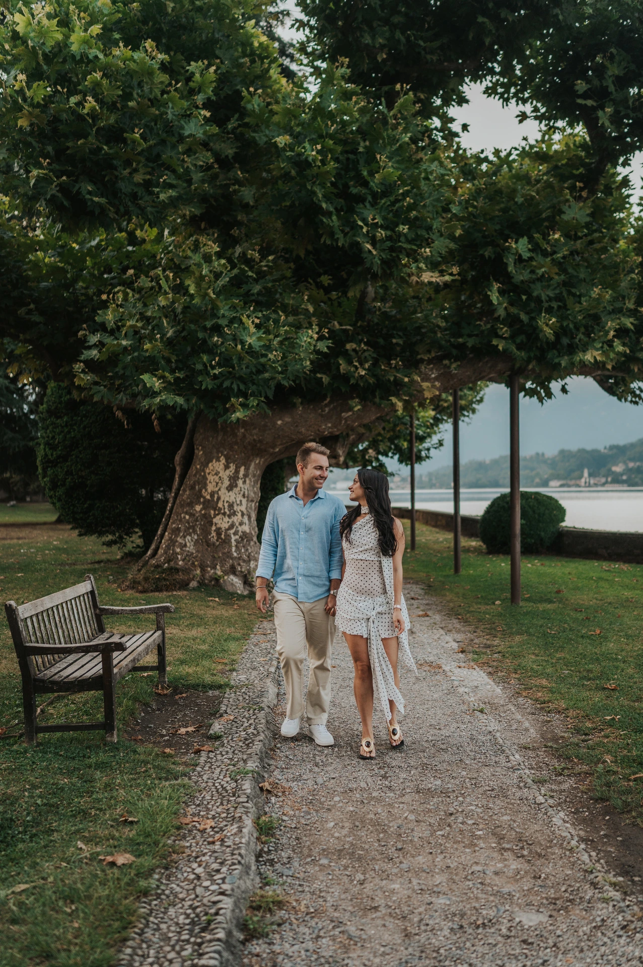 The couple walk hand in hand along a gravel path beneath leafy trees, with a bench and the lake nearby during their Lake Como engagement photoshoot.