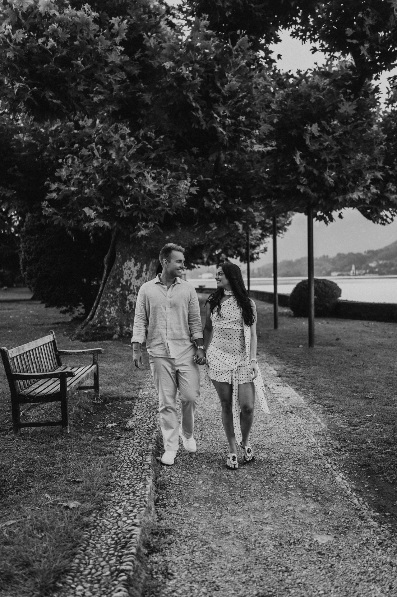 Black-and-white photograph of the couple walking hand in hand beside a bench beneath the park trees during their Lake Como photoshoot.