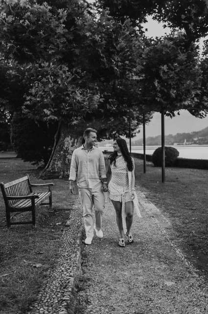 Black-and-white Lake Como photoshoot beneath the park trees Black-and-white photograph of the couple walking hand in hand beside a bench beneath the park trees during their Lake Como photoshoot.