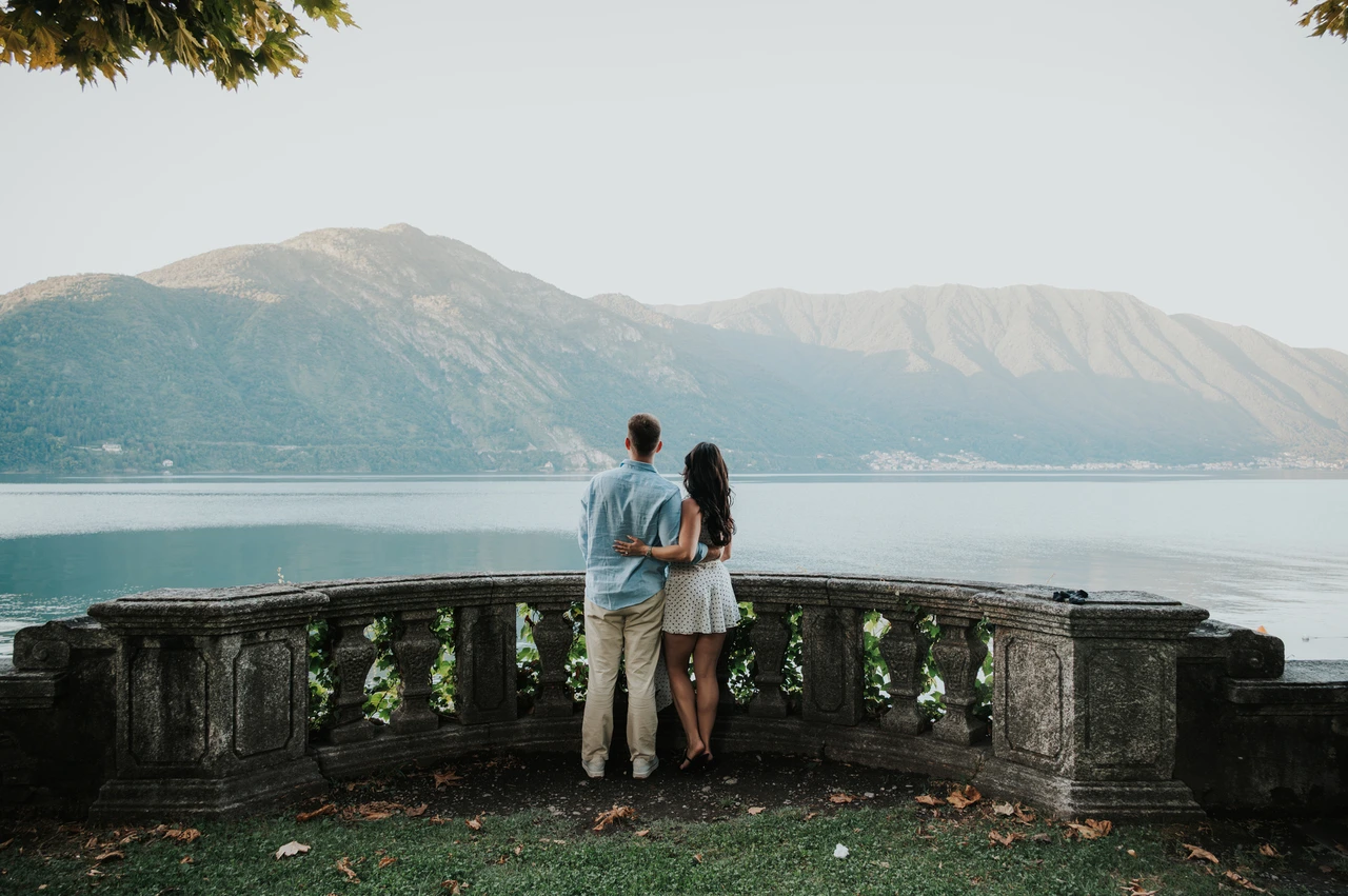 Wide Lake Como engagement photoshoot on the park’s lakeside terrace A wide view of the couple facing the lake from a tree-shaded stone terrace during their Lake Como engagement photoshoot at Parco Civico Teresio Olivelli.