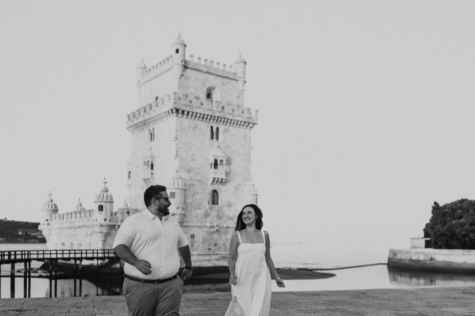 A black-and-white photograph of the couple standing apart and smiling at each other by Belém Tower during their anniversary photoshoot in Lisbon.