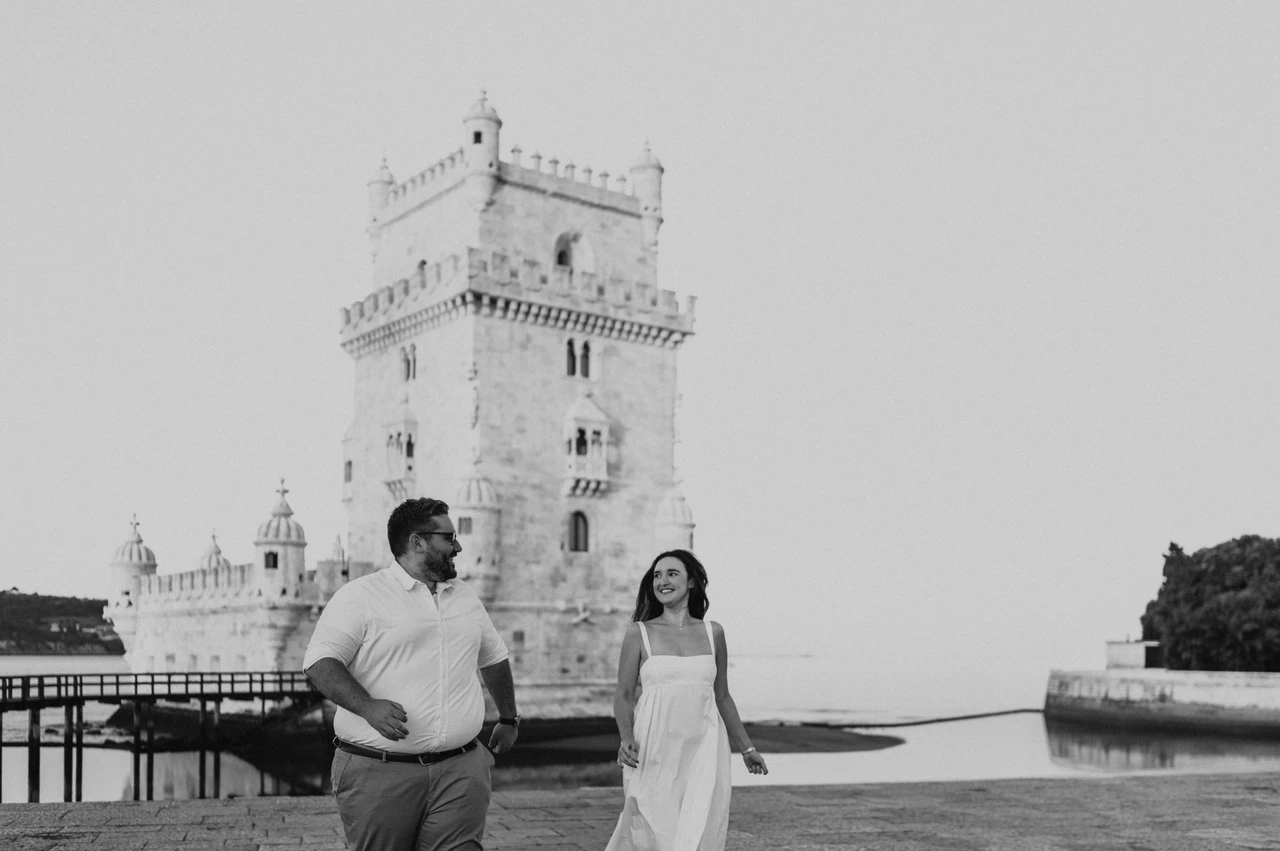 Shared smiles in black and white | Lisbon anniversary photoshoot A black-and-white photograph of the couple standing apart and smiling at each other by Belém Tower during their anniversary photoshoot in Lisbon.