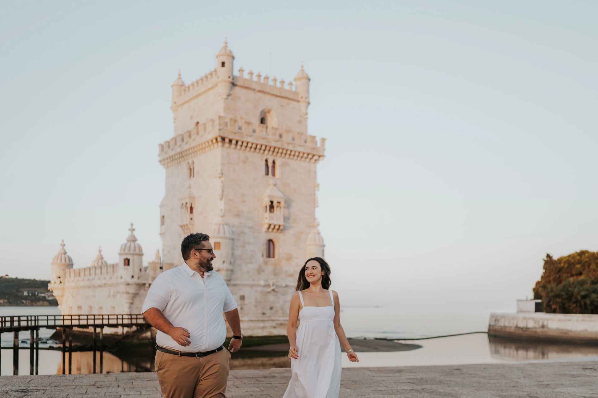 The couple standing apart and smiling at each other beside Belém Tower during their anniversary photoshoot in Lisbon.