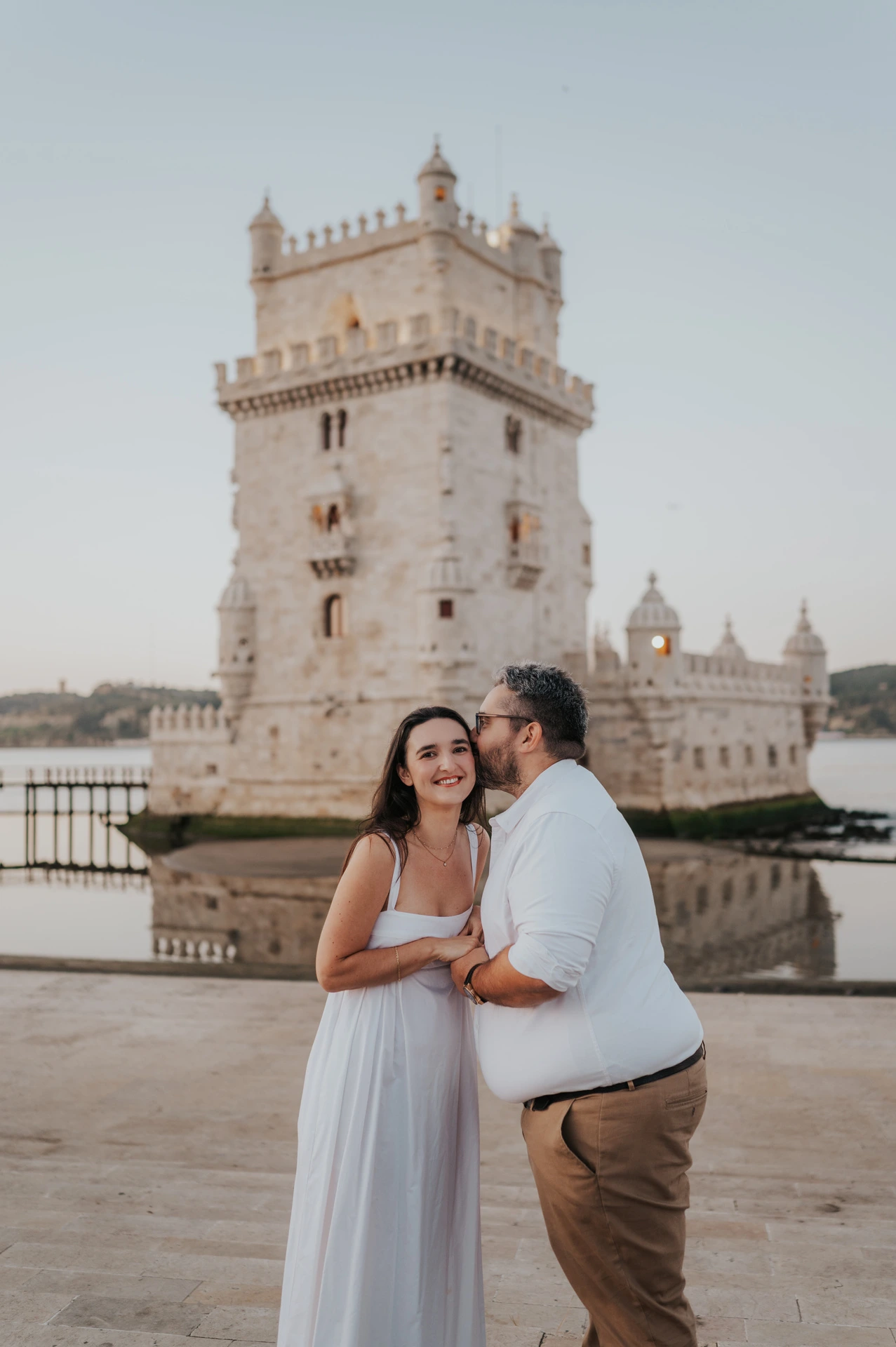 The man kissing his smiling partner on the cheek while they hold hands in front of Belém Tower during their anniversary photoshoot in Lisbon.