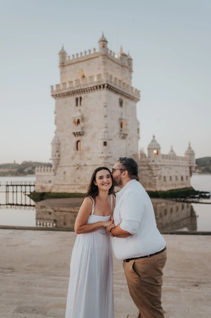The man kissing his smiling partner on the cheek while they hold hands in front of Belém Tower during their anniversary photoshoot in Lisbon.