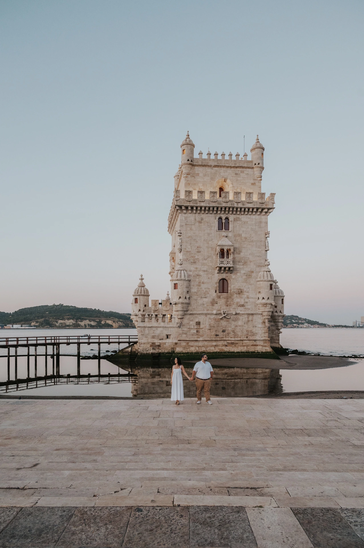 The couple standing together at a distance on the stone paving beside Belém Tower during their anniversary photoshoot in Lisbon.