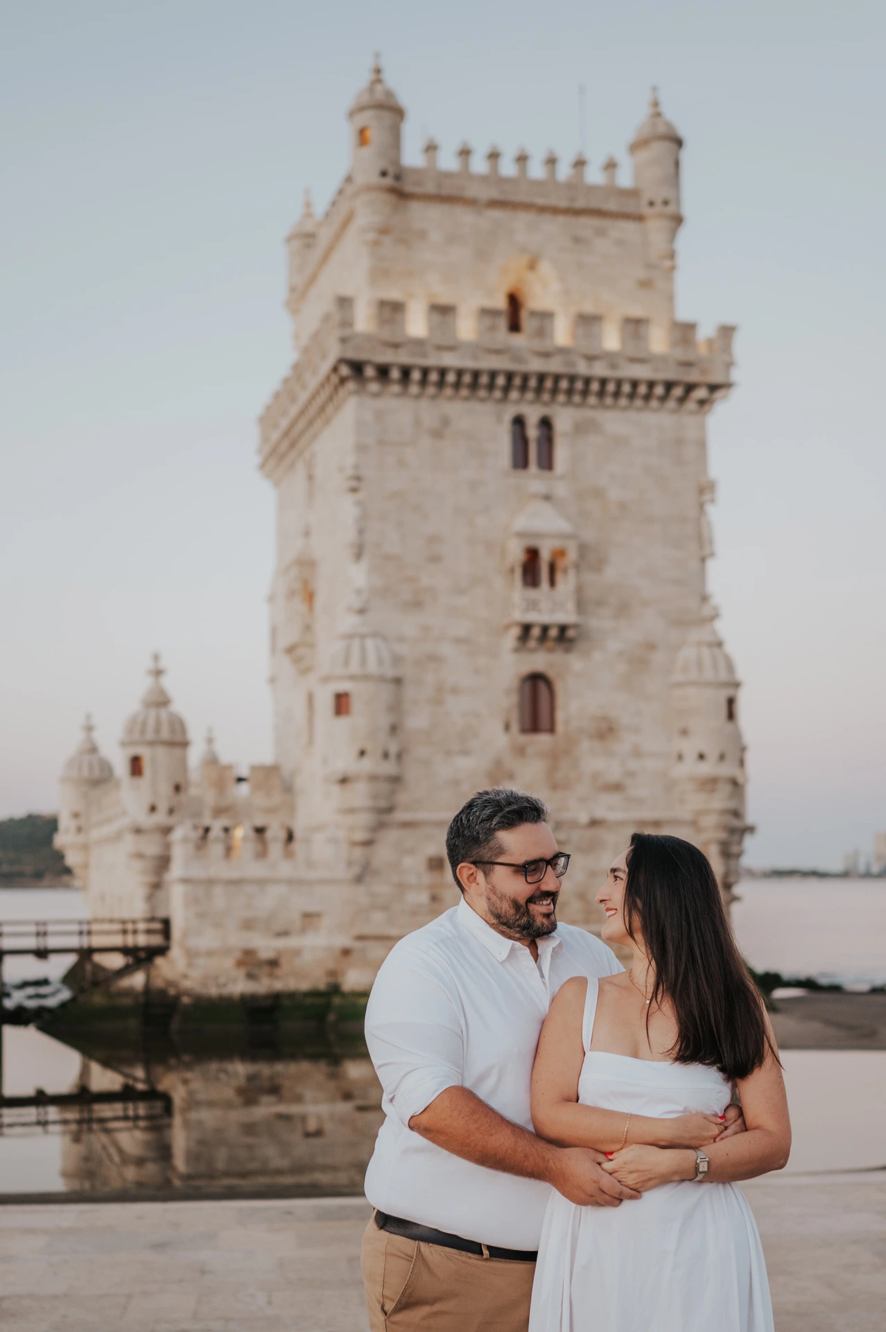 A close portrait of the couple smiling at one another in an embrace with Belém Tower behind them during their anniversary photoshoot in Lisbon.
