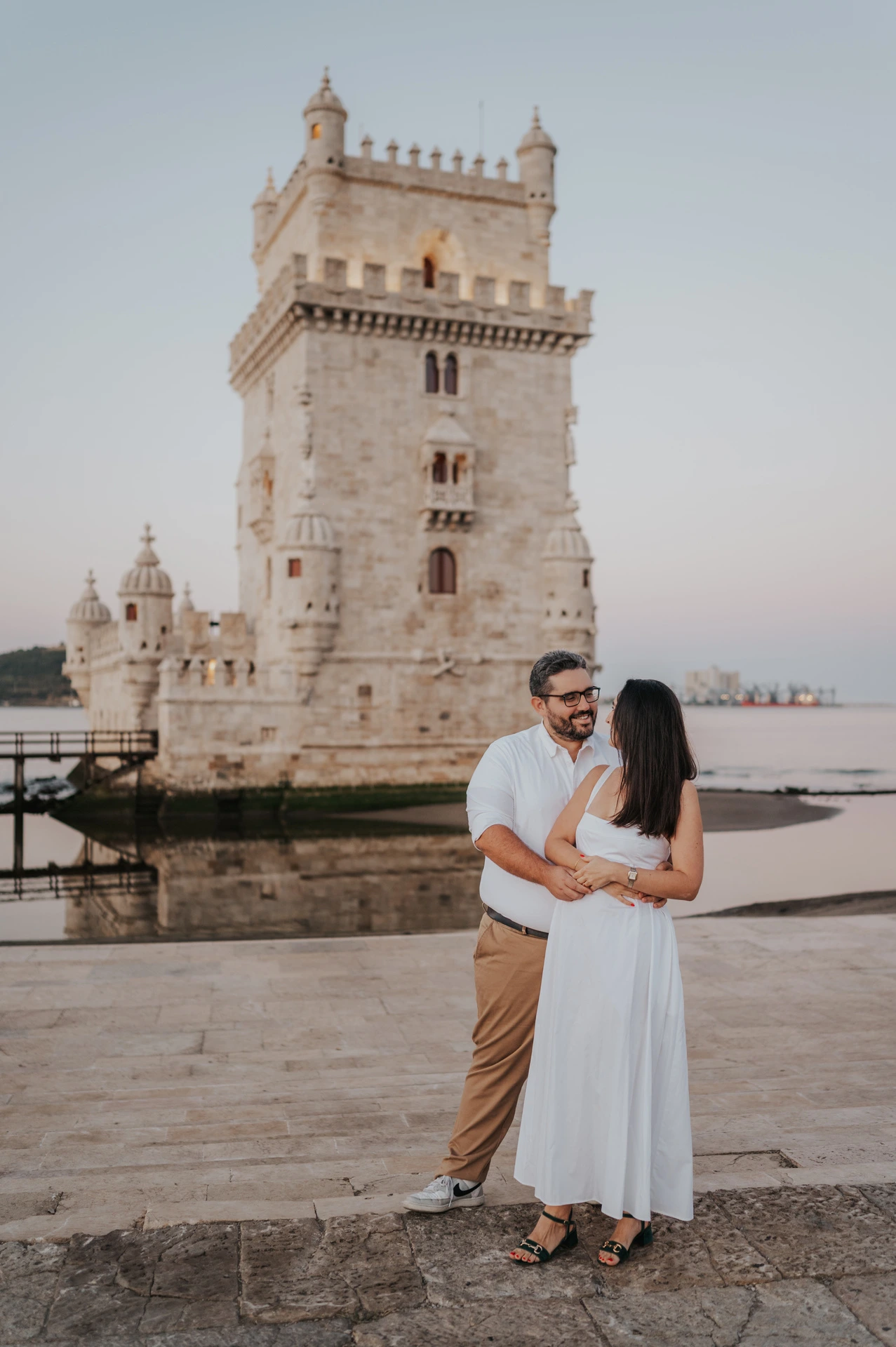 The couple embracing on the paving with Belém Tower behind them during their anniversary photoshoot in Lisbon.