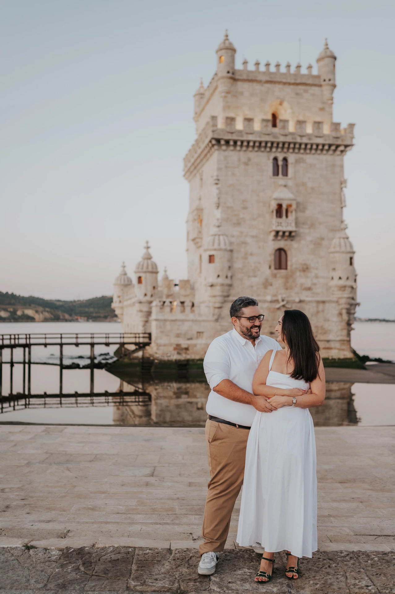The couple smiling at each other while embracing on the paving by Belém Tower during their anniversary photoshoot in Lisbon.