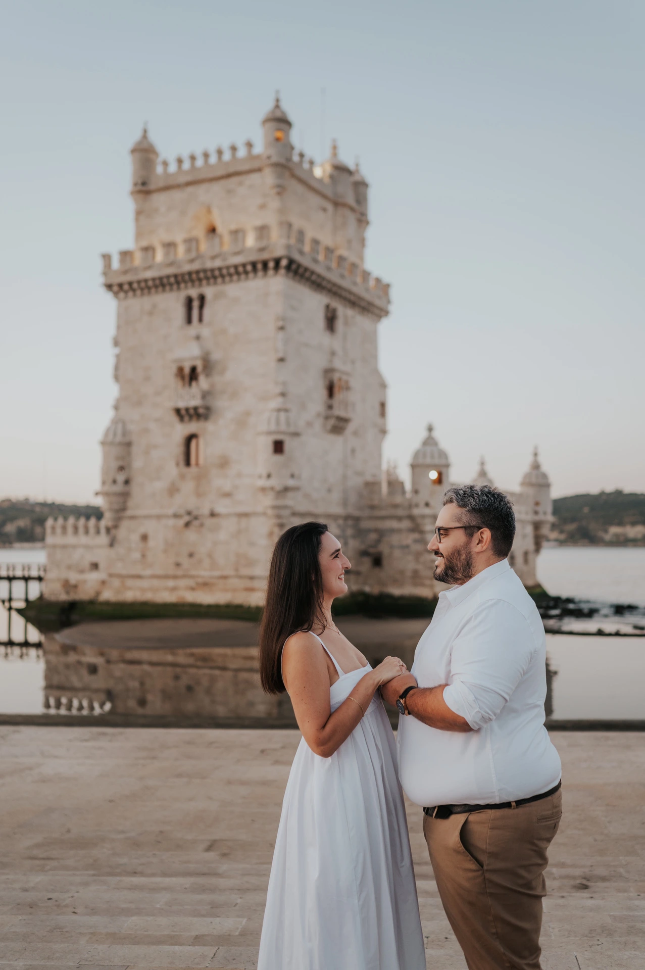 The couple facing each other and holding hands with Belém Tower in the background during their anniversary photoshoot in Lisbon.