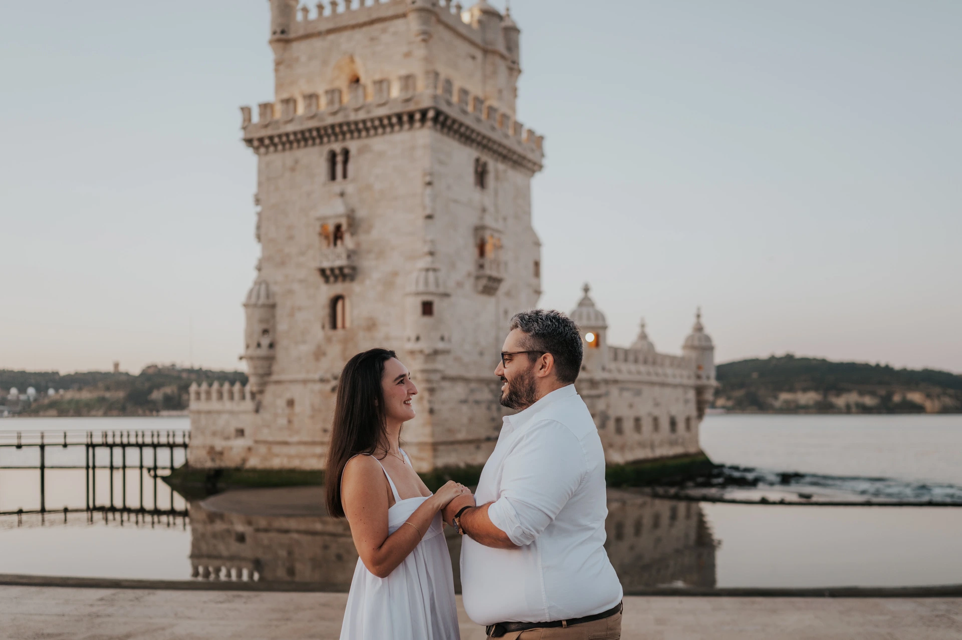 A landscape portrait of the couple holding hands and looking at each other beside Belém Tower during their anniversary photoshoot in Lisbon.