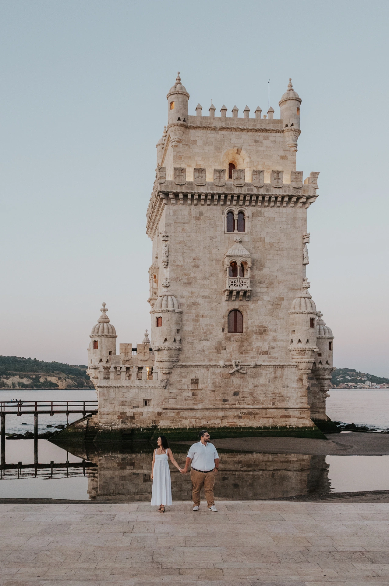 A full view of Belém Tower with the couple standing hand in hand in the foreground during their anniversary photoshoot in Lisbon.