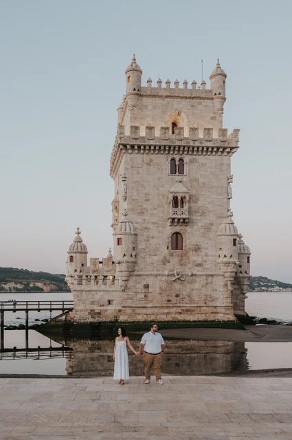 A full view of Belém Tower with the couple standing hand in hand in the foreground during their anniversary photoshoot in Lisbon.