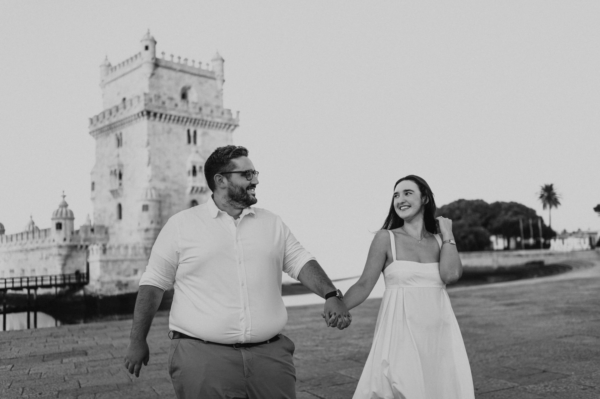 A black-and-white landscape photograph of the couple smiling at each other as they walk by Belém Tower during their anniversary photoshoot in Lisbon.