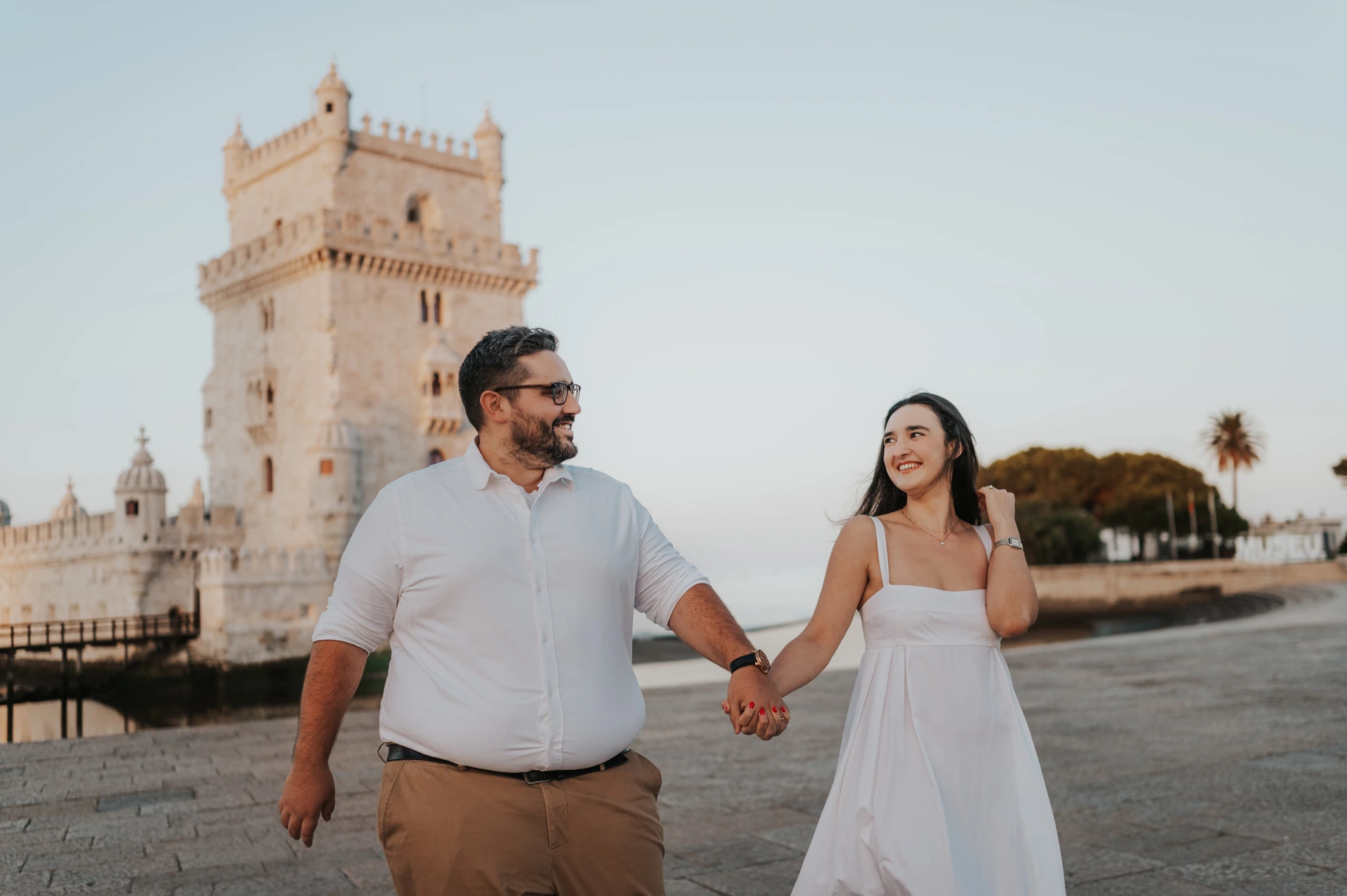 A closer landscape view of the couple smiling at each other while walking hand in hand by Belém Tower during their anniversary photoshoot in Lisbon.