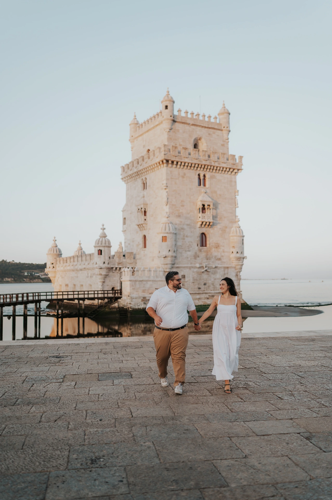 A full-length portrait of the couple walking together beside Belém Tower during their anniversary photoshoot in Lisbon.
