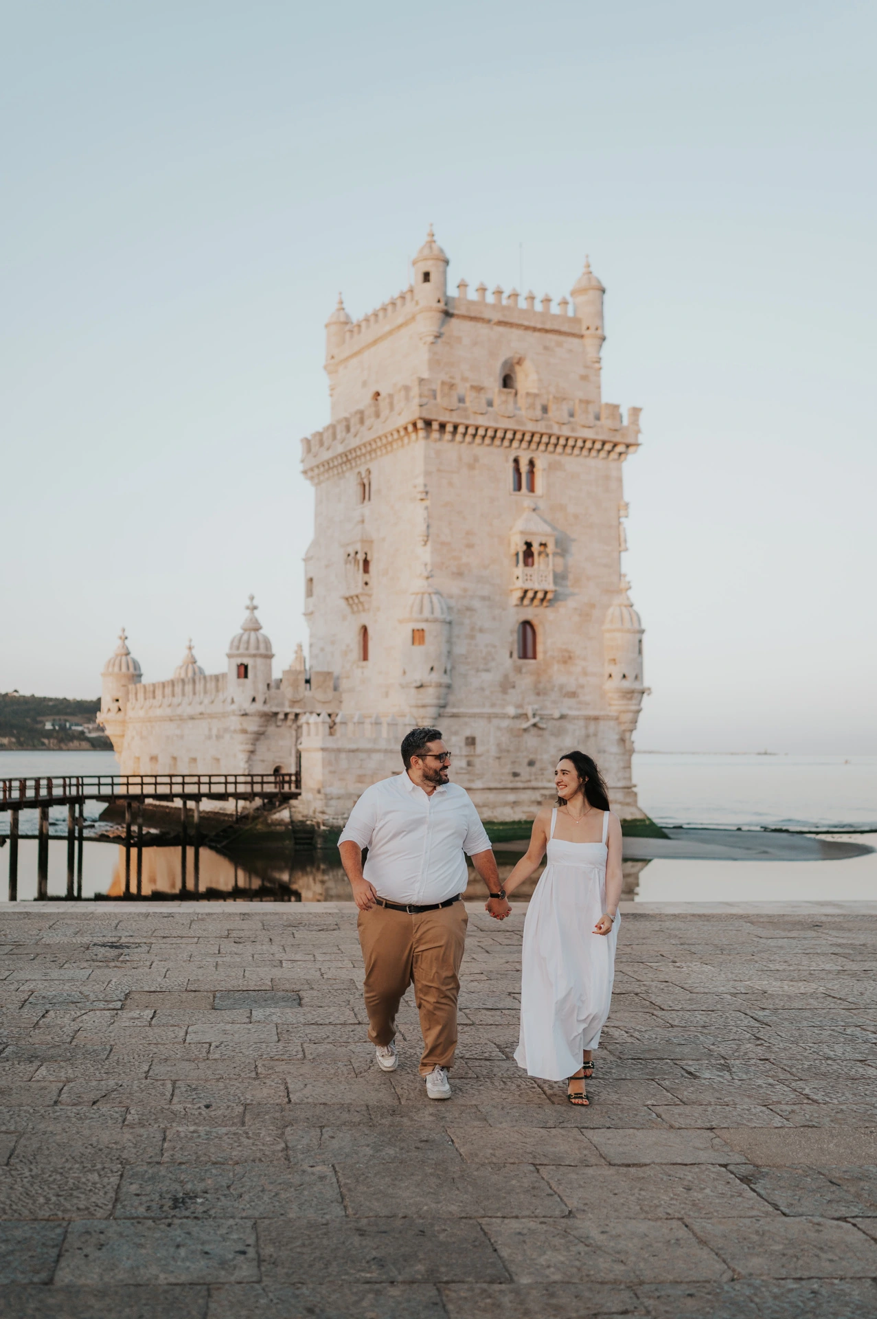 The couple walking hand in hand across the open paving with Belém Tower in the background during their anniversary photoshoot in Lisbon.