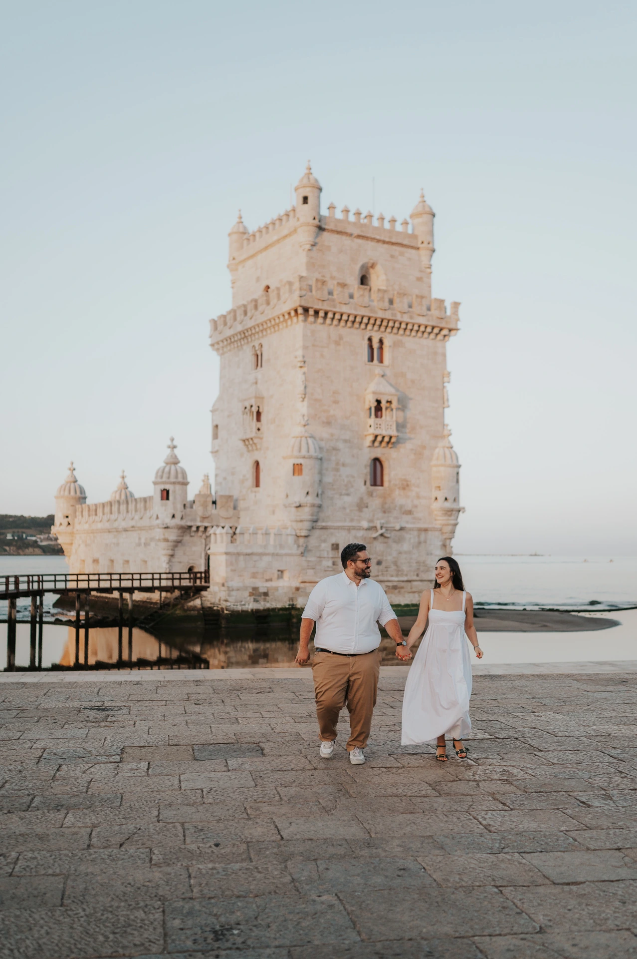The couple looking at each other while walking hand in hand in front of Belém Tower during their anniversary photoshoot in Lisbon.