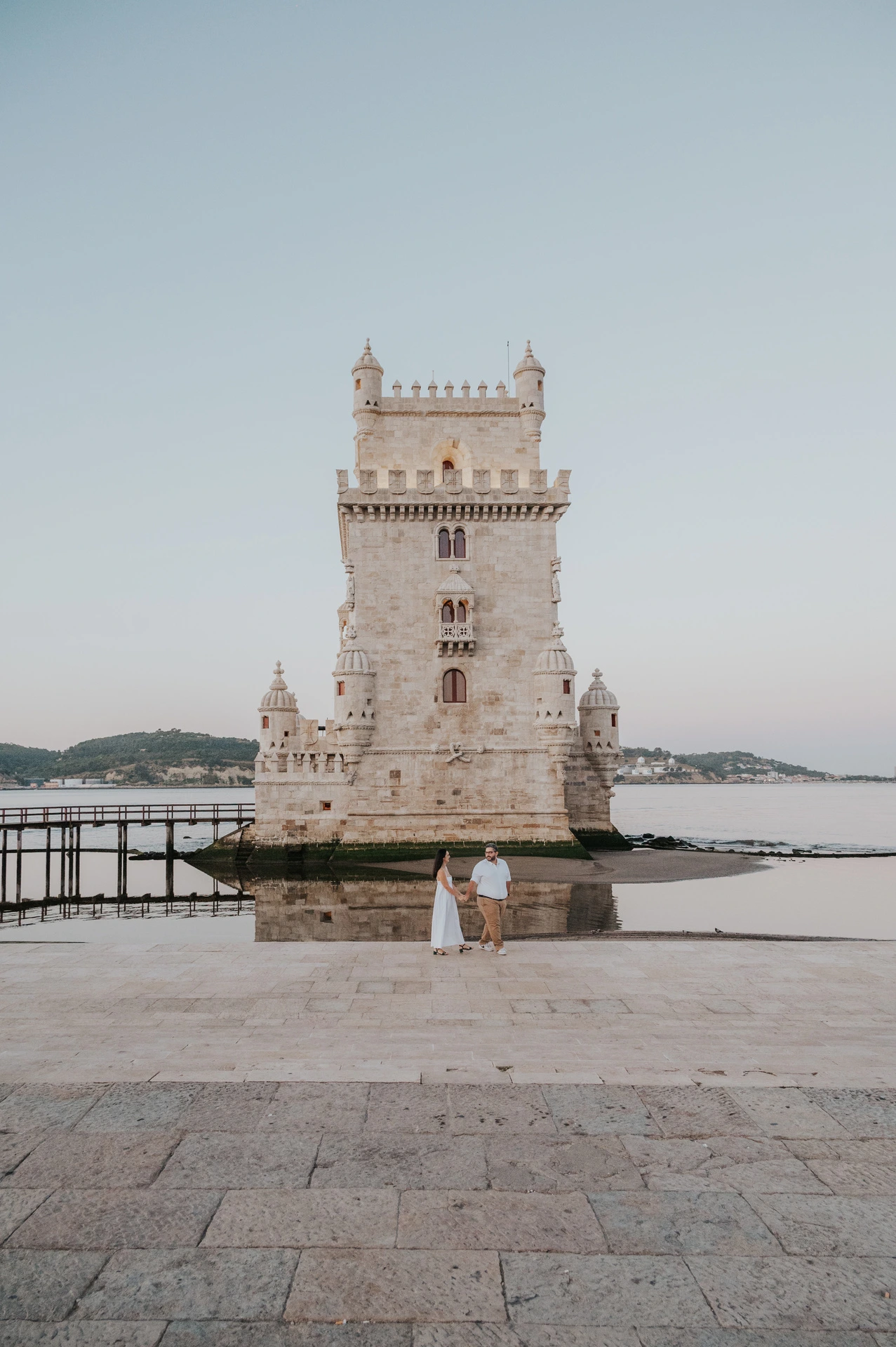 The couple standing together in front of Belém Tower above a broad stretch of stone paving during their anniversary photoshoot in Lisbon.