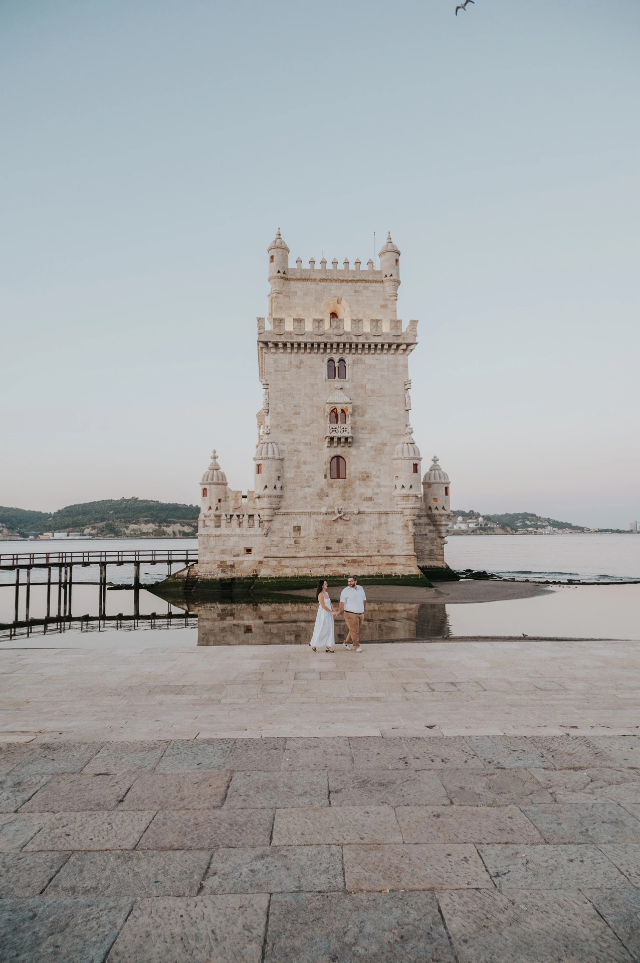 A wide vertical view of the couple standing close together with Belém Tower beyond the open paving during their anniversary photoshoot in Lisbon.