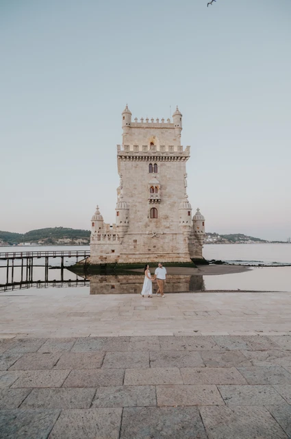 A quiet portrait across the open paving | Lisbon anniversary photoshoot A wide vertical view of the couple standing close together with Belém Tower beyond the open paving during their anniversary photoshoot in Lisbon.