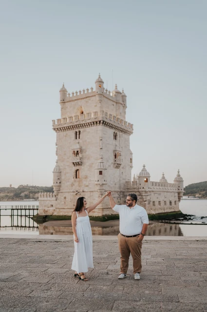 A playful hand-in-hand moment | Lisbon anniversary photoshoot A wider portrait of the couple holding their joined hands up with Belém Tower behind them during their anniversary photoshoot in Lisbon.