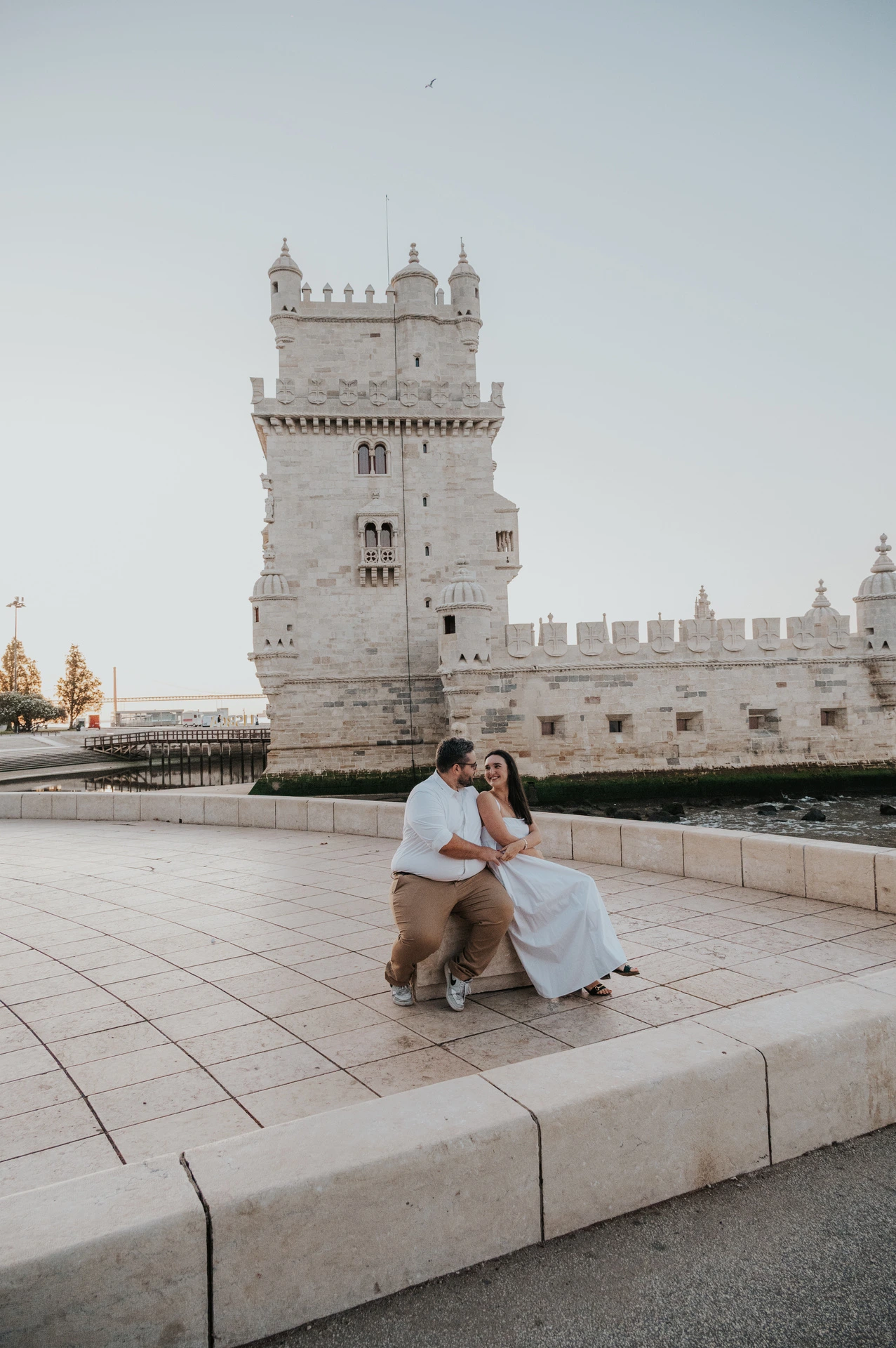 The couple sitting close together on the waterfront ledge in front of Belém Tower during their anniversary photoshoot in Lisbon.