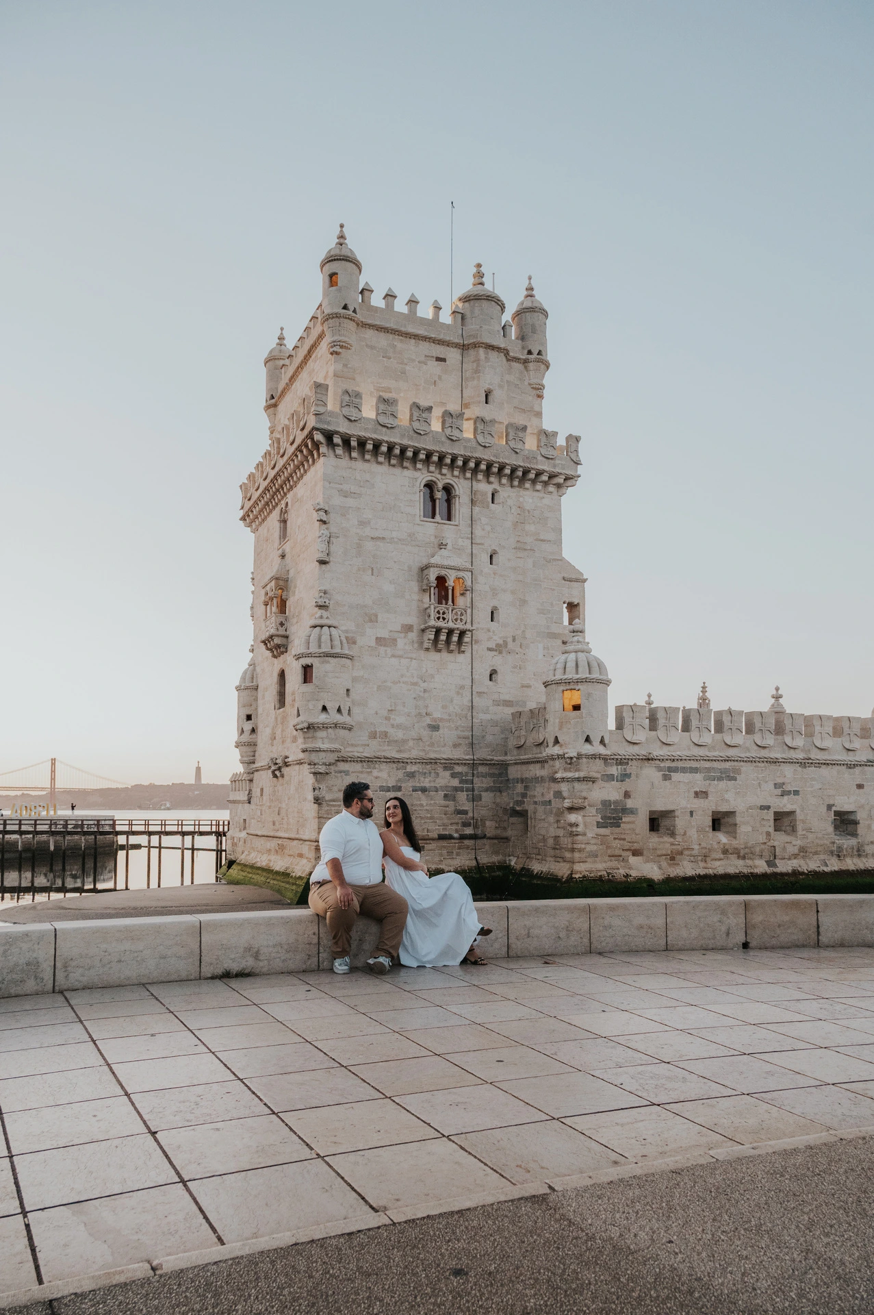 The couple sitting together on a low ledge with open paving and Belém Tower around them during their anniversary photoshoot in Lisbon.