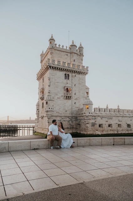 A seated portrait across the open paving | Lisbon anniversary photoshoot The couple sitting together on a low ledge with open paving and Belém Tower around them during their anniversary photoshoot in Lisbon.