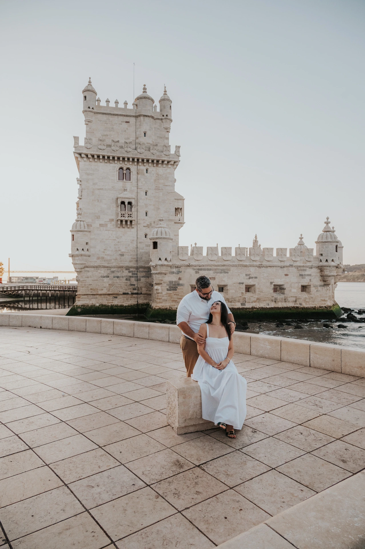 The woman seated on the low ledge with her partner leaning towards her beside Belém Tower during their anniversary photoshoot in Lisbon.