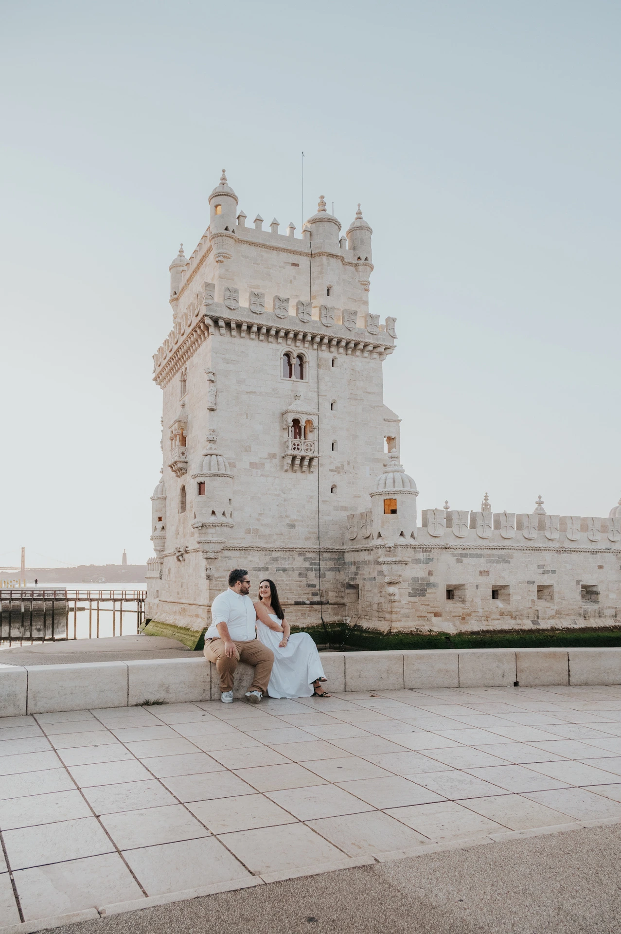 A distant view of the couple sitting close together on a low stone ledge beside Belém Tower during their anniversary photoshoot in Lisbon.