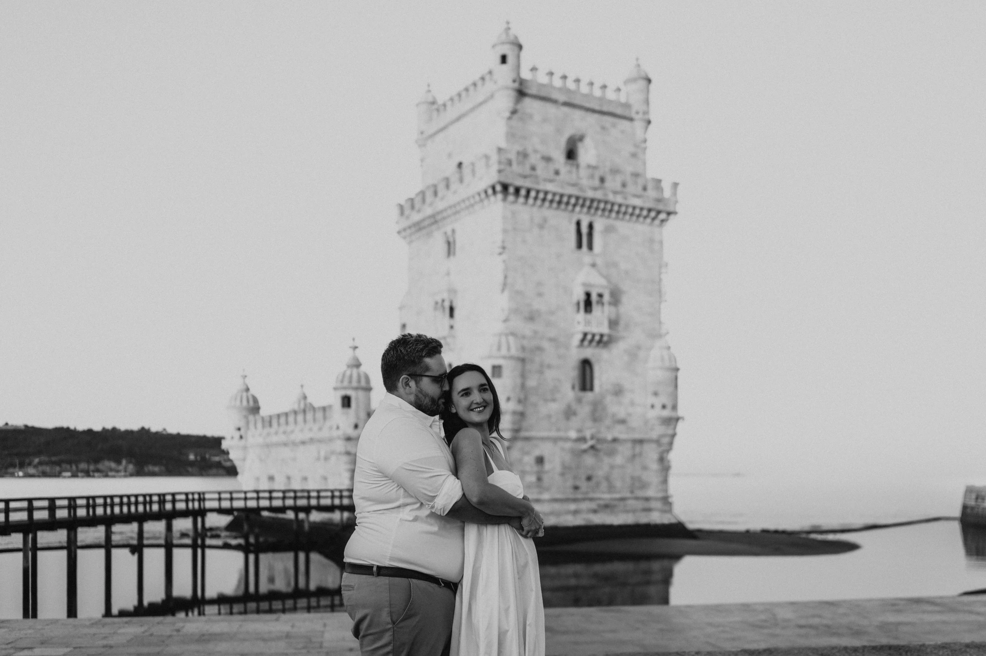 A black-and-white landscape portrait of the couple embracing with Belém Tower behind them during their anniversary photoshoot in Lisbon.