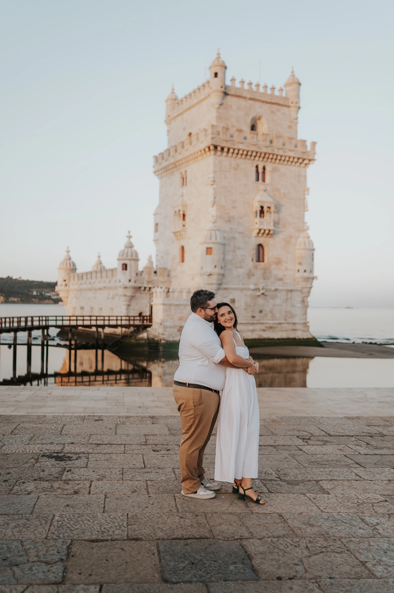 A full-length portrait of the couple in a close side embrace beside Belém Tower during their anniversary photoshoot in Lisbon.