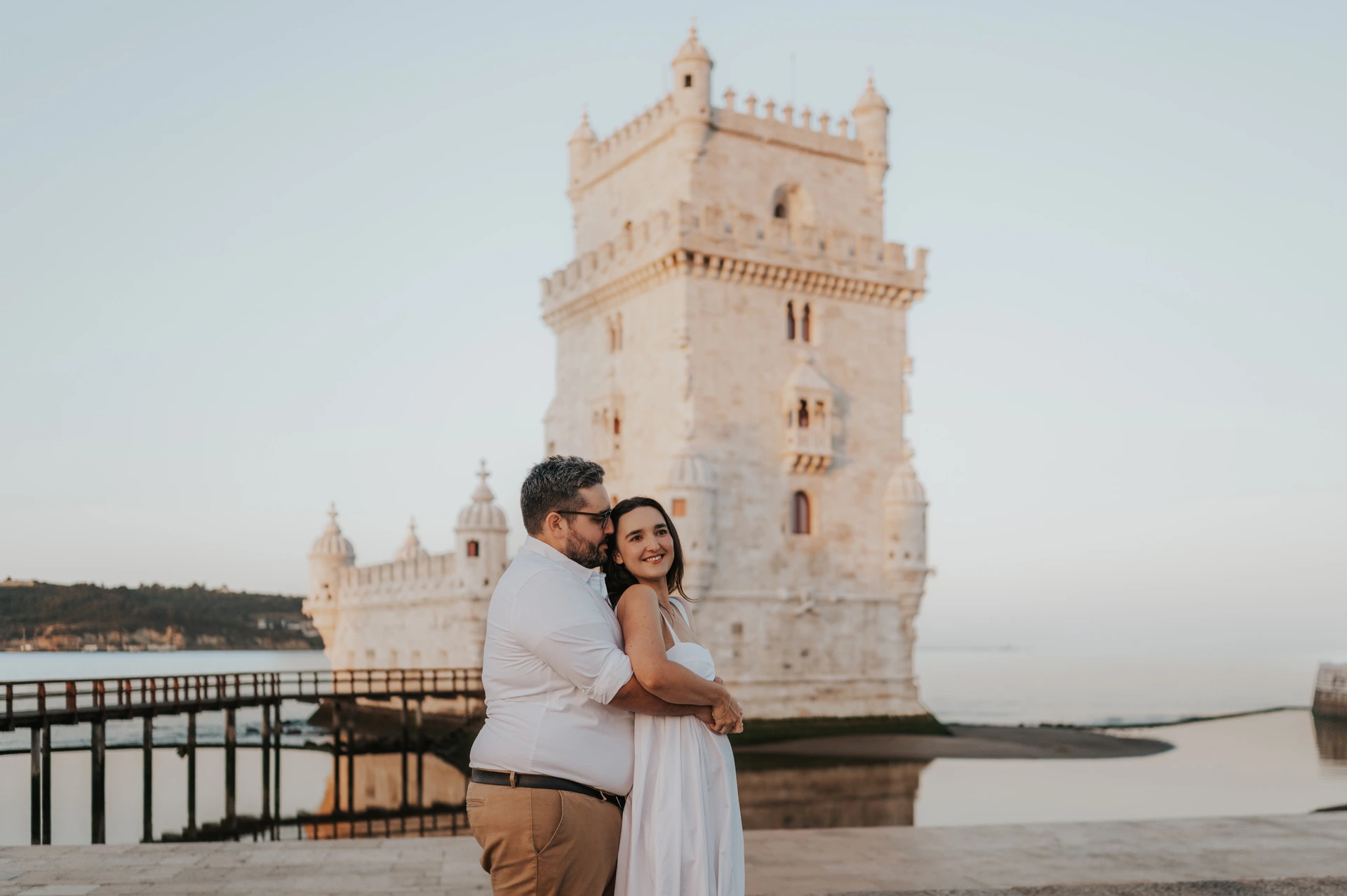 The man kissing his smiling partner near her temple while embracing her in front of Belém Tower during their anniversary photoshoot in Lisbon.