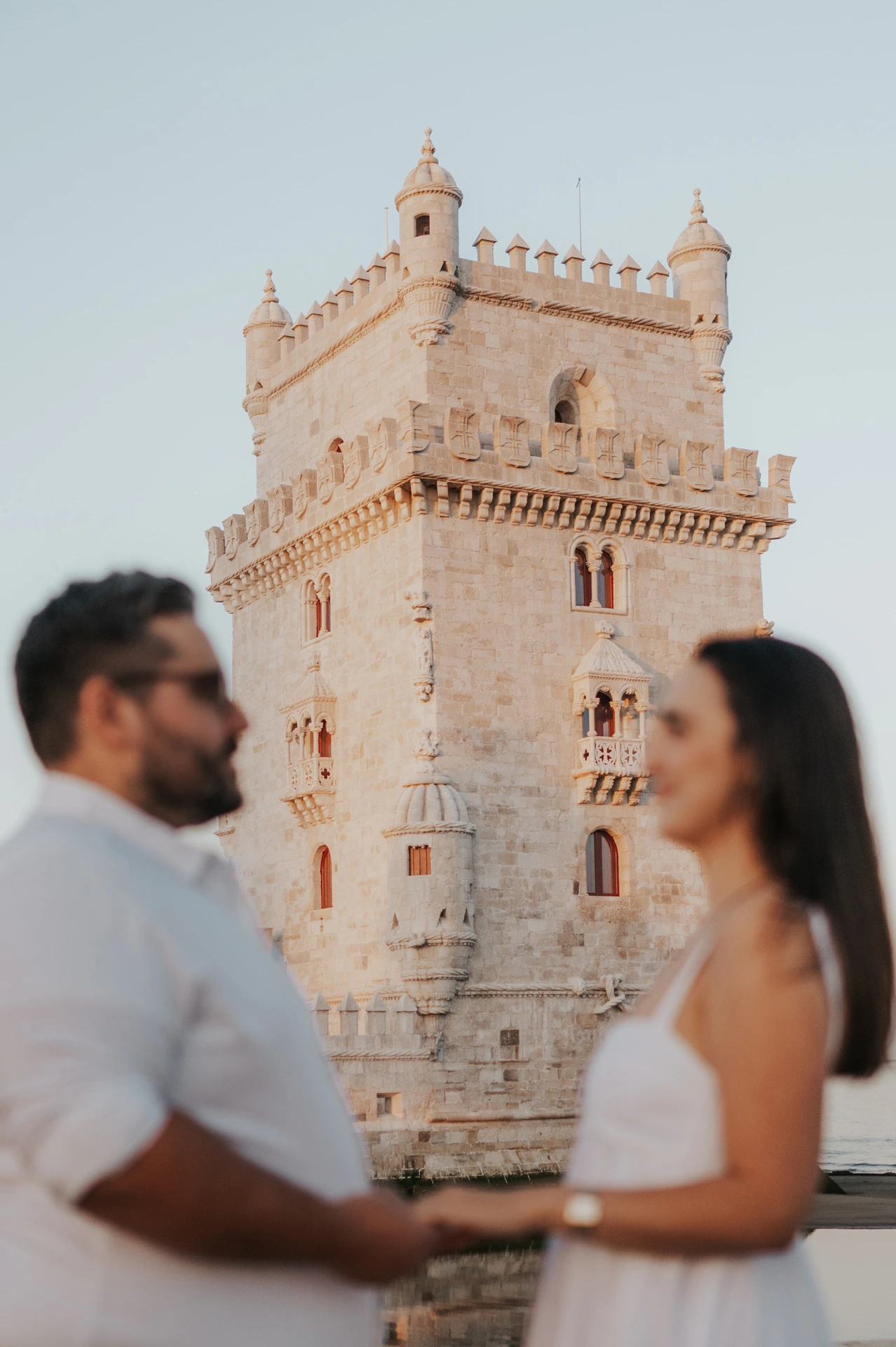 A softly focused portrait of the couple holding hands with Belém Tower sharply visible behind them during their anniversary photoshoot in Lisbon.