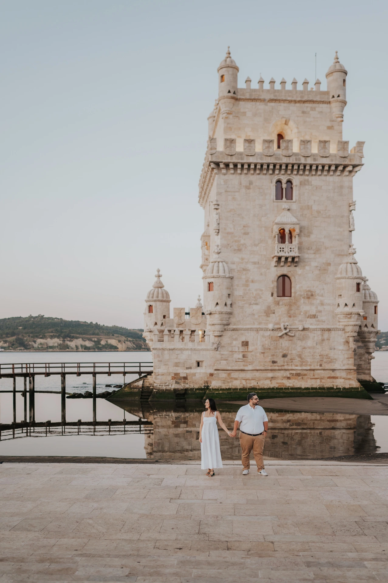 The couple standing hand in hand at a distance in front of Belém Tower during their anniversary photoshoot in Lisbon.