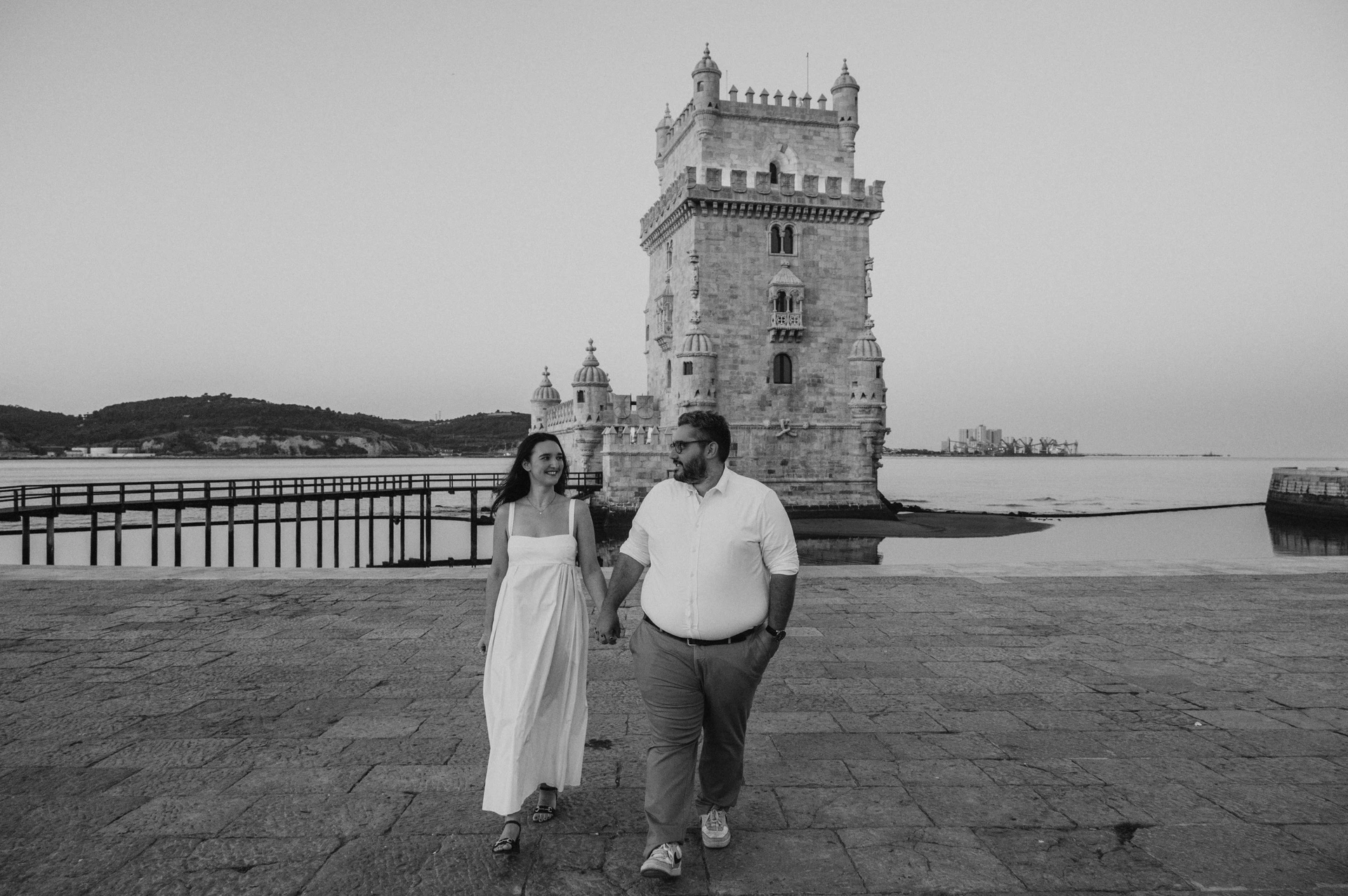 A black-and-white photograph of the couple walking together in front of Belém Tower during their anniversary photoshoot in Lisbon.
