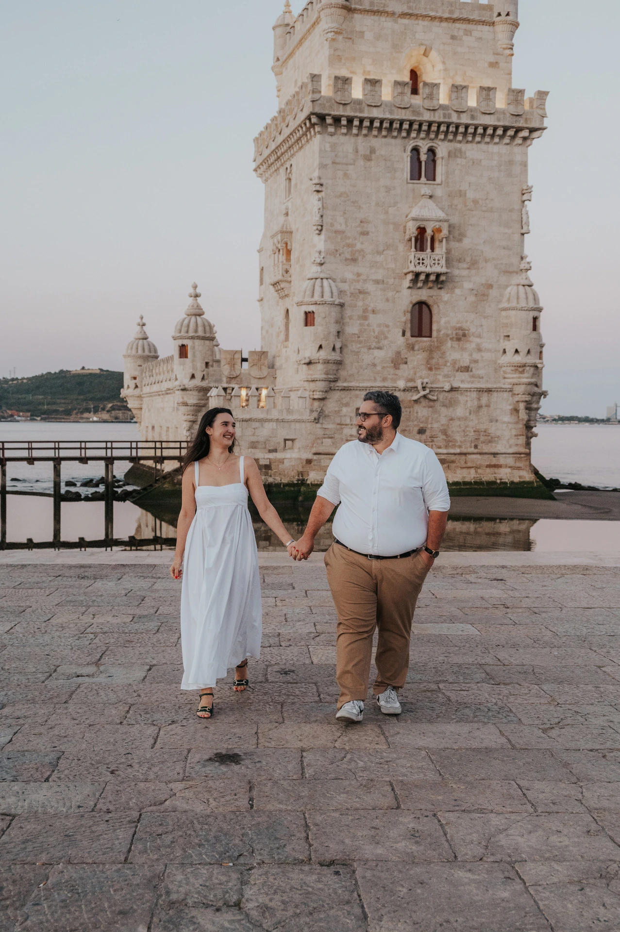 The couple looking at each other as they walk hand in hand with Belém Tower behind them during their anniversary photoshoot in Lisbon.