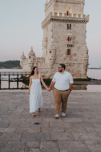 A shared glance on their morning walk | Lisbon anniversary photoshoot The couple looking at each other as they walk hand in hand with Belém Tower behind them during their anniversary photoshoot in Lisbon.