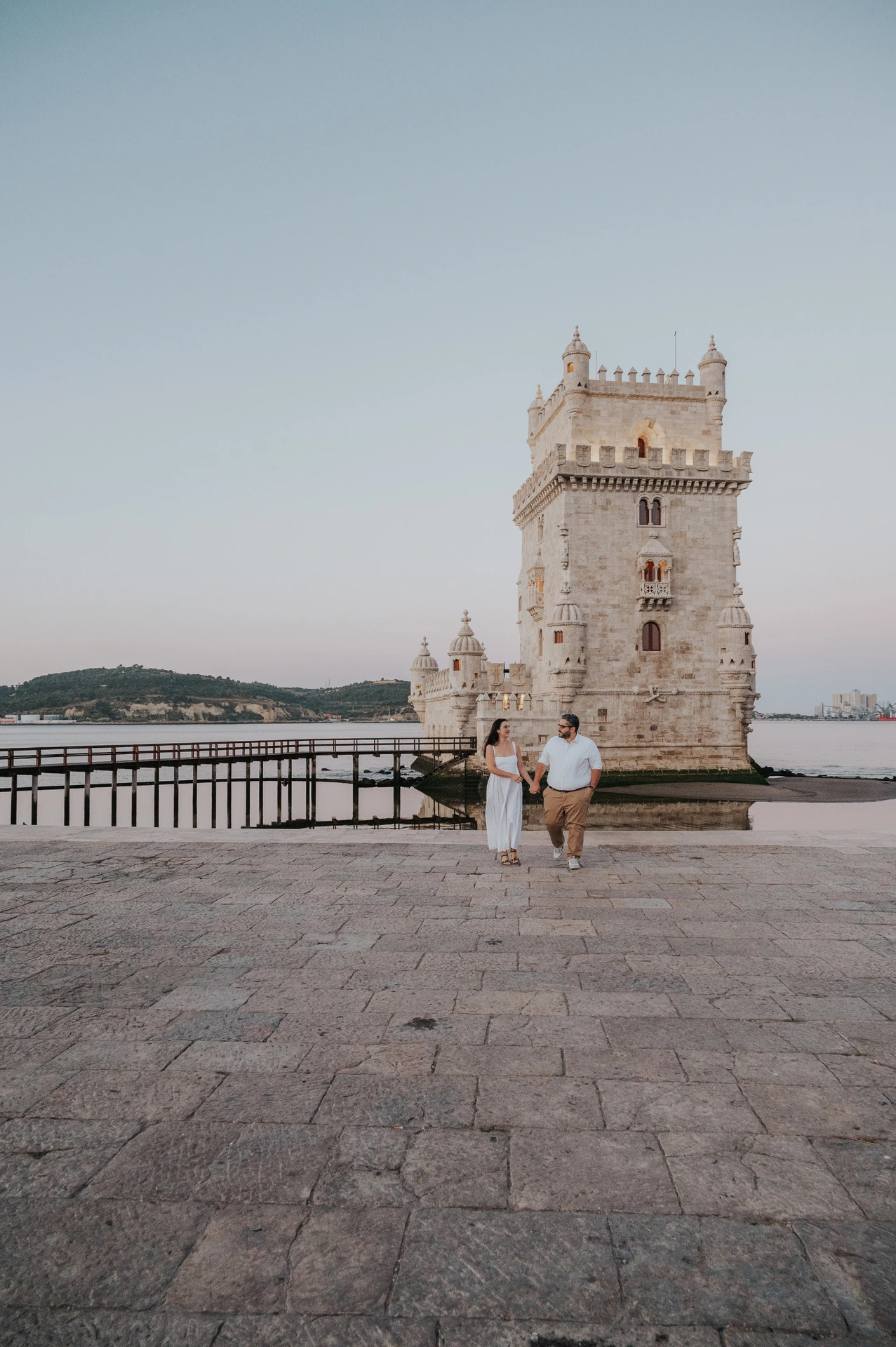 A distant view of the couple walking hand in hand on the paving in front of Belém Tower during their anniversary photoshoot in Lisbon.