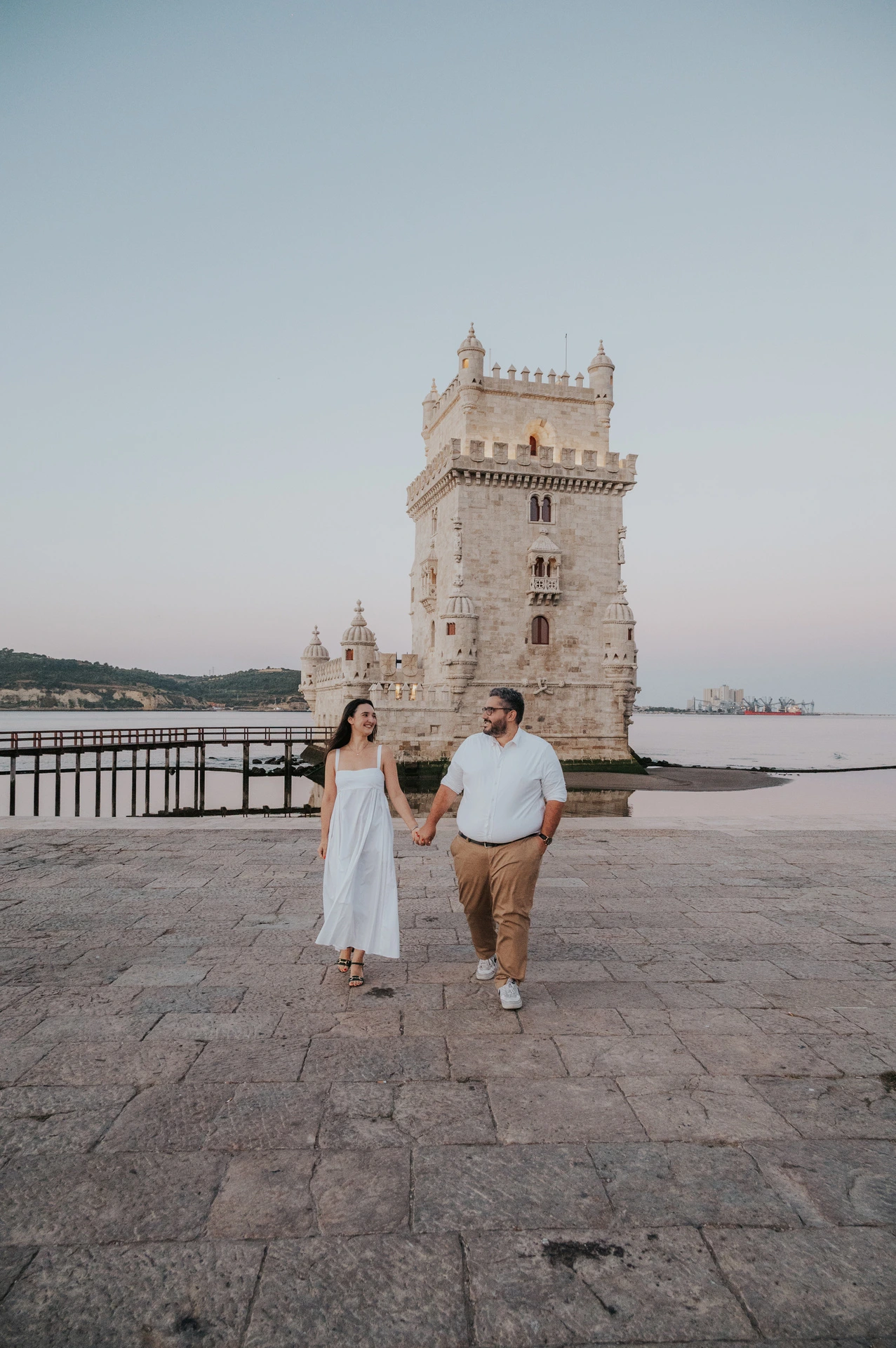 A full-length view of the couple walking hand in hand across the stone paving by Belém Tower during their anniversary photoshoot in Lisbon.