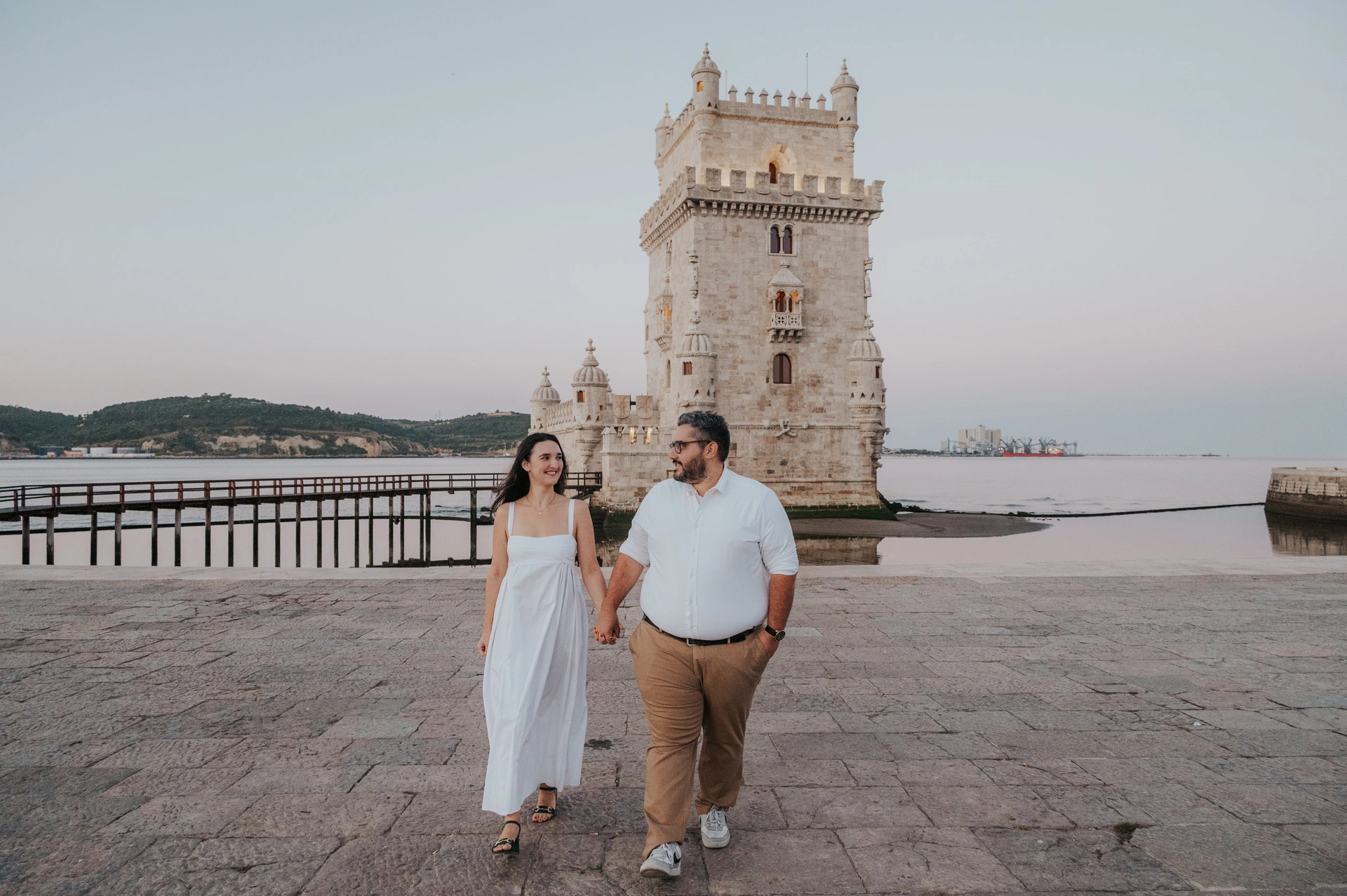A landscape photograph of the couple walking together with Belém Tower and the water behind them during their anniversary photoshoot in Lisbon.