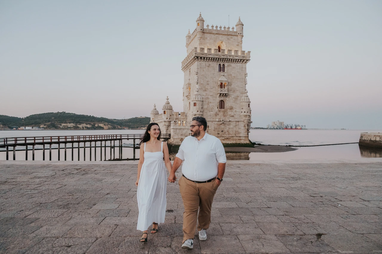 A morning walk beside Belém Tower | Lisbon anniversary photoshoot A landscape photograph of the couple walking together with Belém Tower and the water behind them during their anniversary photoshoot in Lisbon.