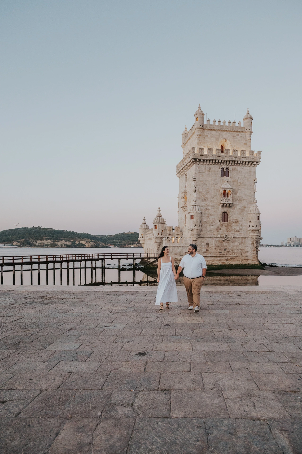 The couple walking towards the camera beneath an open sky beside Belém Tower during their anniversary photoshoot in Lisbon.