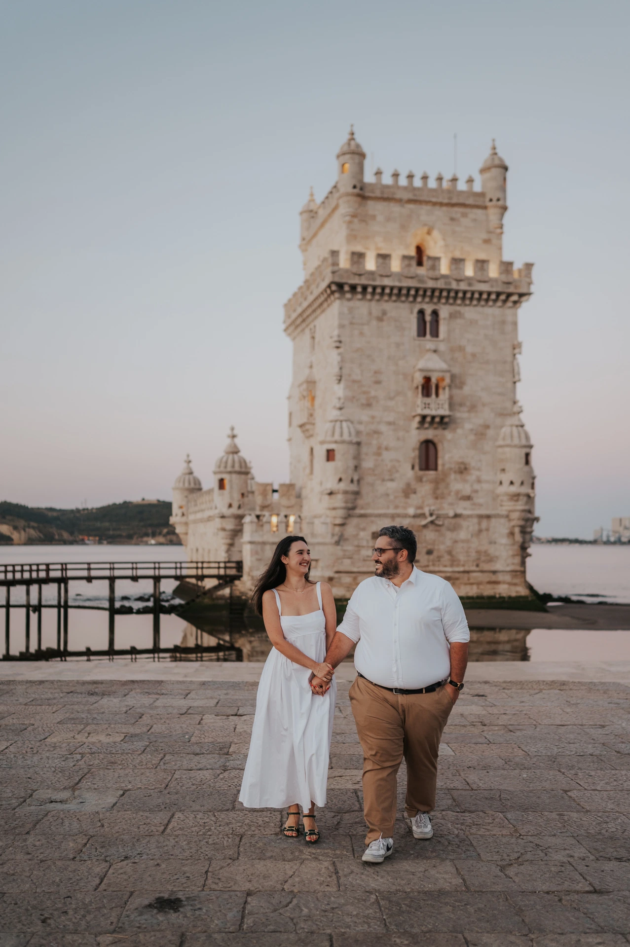 The couple sharing a glance as they walk beside the waterfront railings at Belém Tower during their anniversary photoshoot in Lisbon.