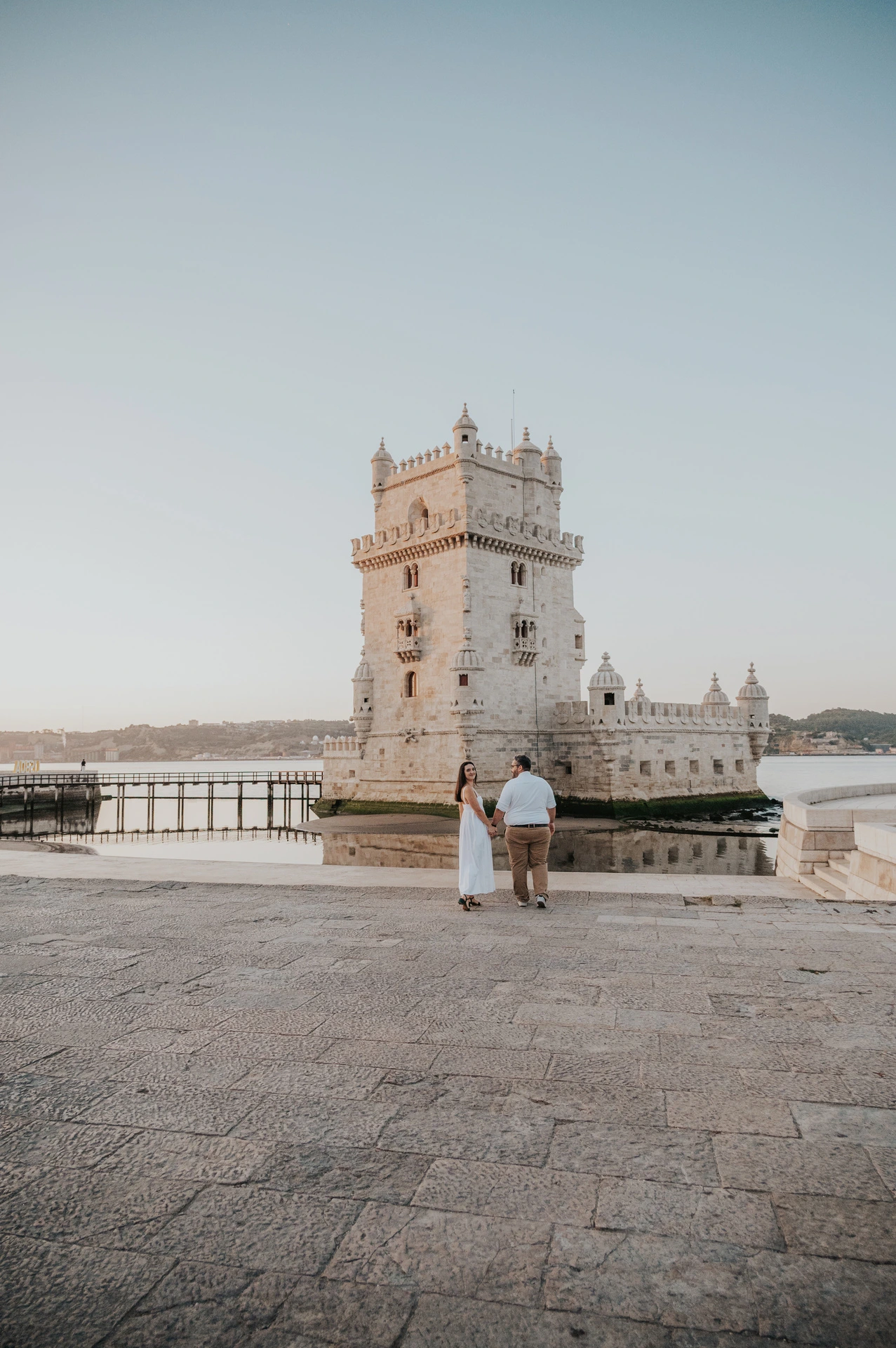 A wide view of the couple walking on the stone paving with Belém Tower and the water behind them during their anniversary photoshoot in Lisbon.